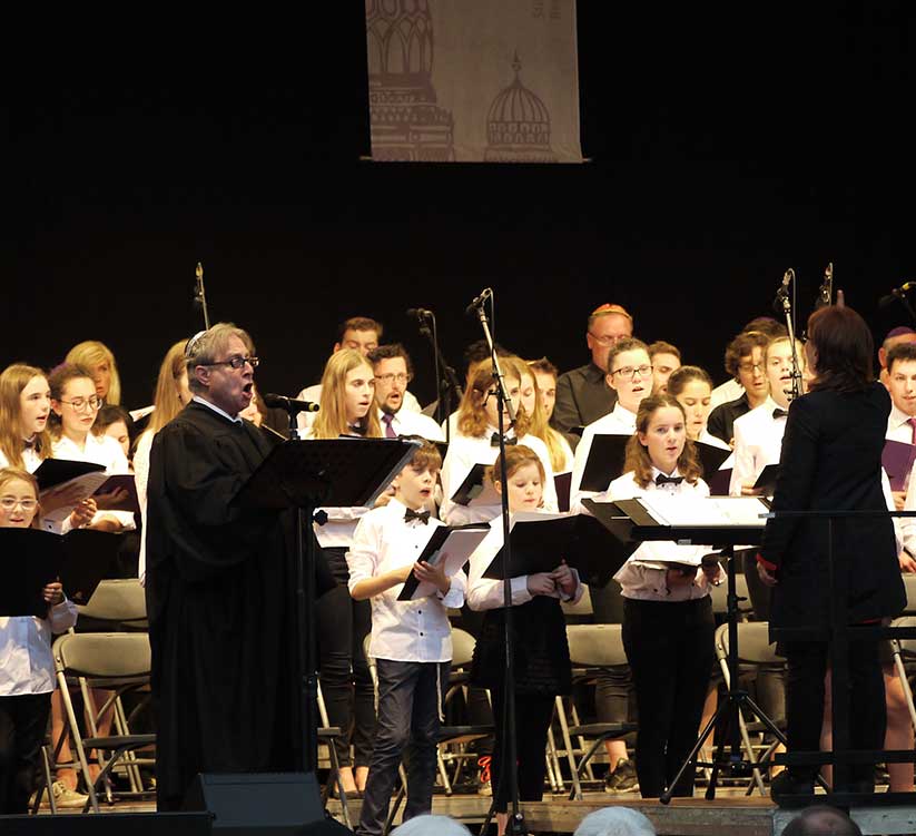 Der Synagogenchor singt gemeinsam ein Konzert auf einer Bühne. Dirigiert werden sie von Regina Yantian (unten rechts im Bild) und begleitet vom Kantor Isaac Sheffer (unten links im Bild).