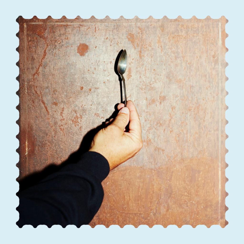 Quadratisches Bild, auf dem eine Hand einen Silberlöffel hält. Das Bild hat einen ausgestanzten Rand wie bei Briefmarken und ist umrandet von einem hellblauen Rahmen.