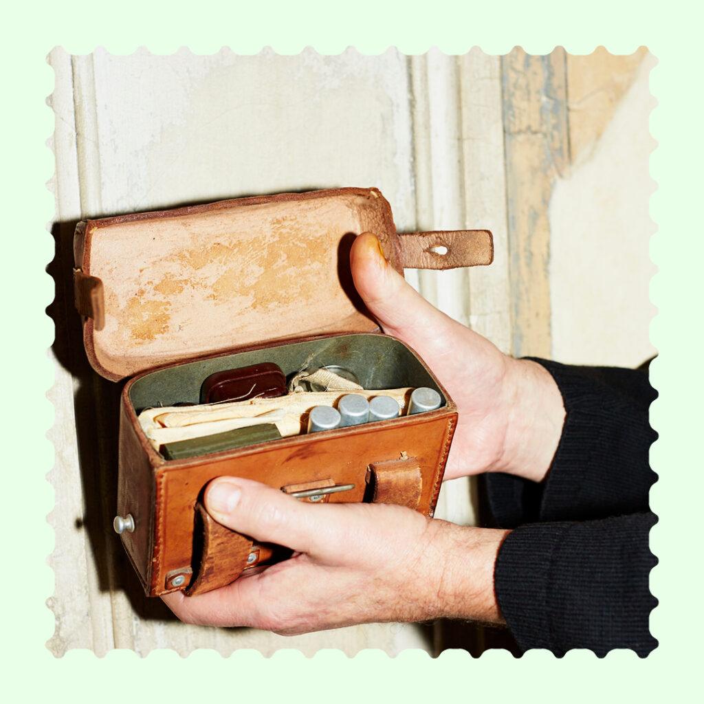 Quadratisches Bild, auf dem Hände ein Medizintäschchen halten. Das Bild hat einen ausgestanzten Rand wie bei Briefmarken und ist umrandet von einem hellgrünen Rahmen.