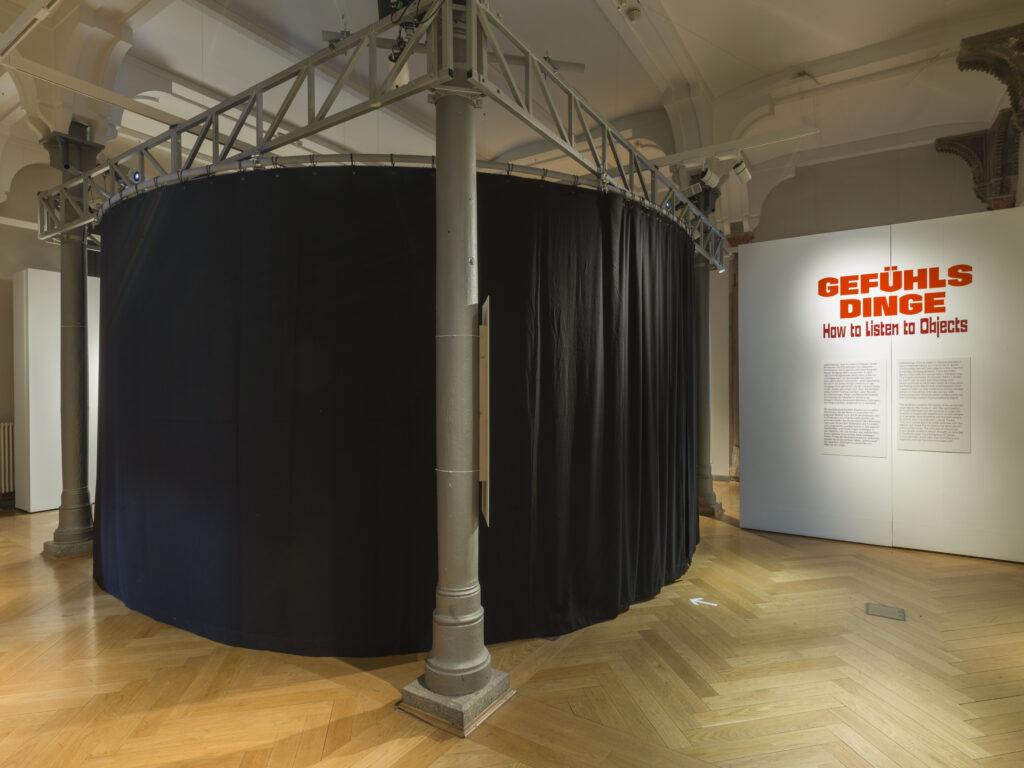 Blick in die Wechselausstellung Gefühlsdinge - How to Listen to Objects: Eingangsbereich mit weißer Wand, die den Titel in roter Schrift und die Einleitung zeigt (rechts), links ist ein raumfüllendes, rundes Filmzelt von außen sichtbar (schwarzer Stoff), davor eine Säule mit Erklärtext.
