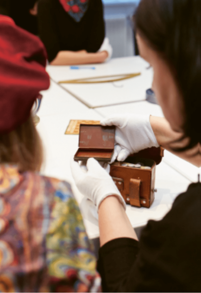 Foto, das zwei Personen von hinten zeigt, sie sitzen an einem weißen Tisch, die rechte Person trägt weiße Handschuhe und hält ein Zigaretten-Etui in den Händen. Daneben steht ein ledernes Medizintäschchen.